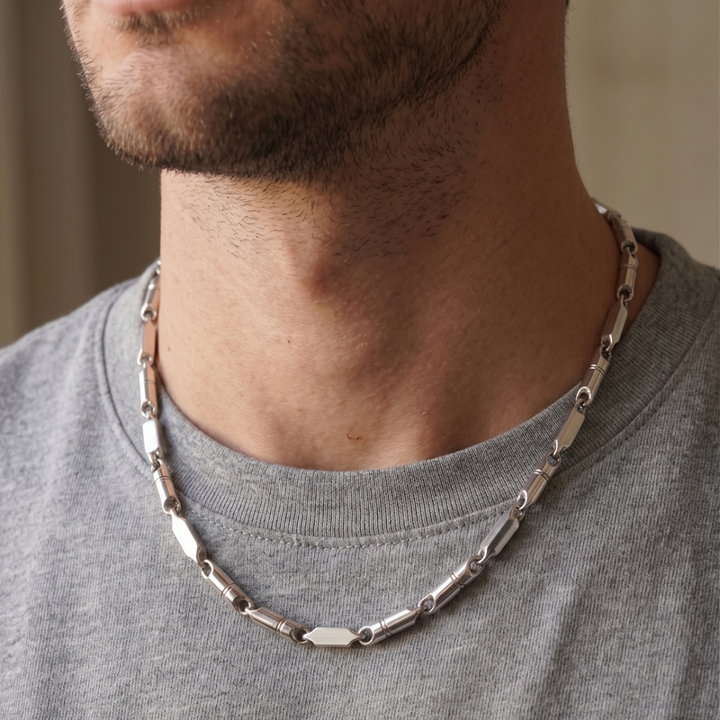 A close-up of a man wearing a silver-toned geometric link chain necklace over a gray crewneck t-shirt.