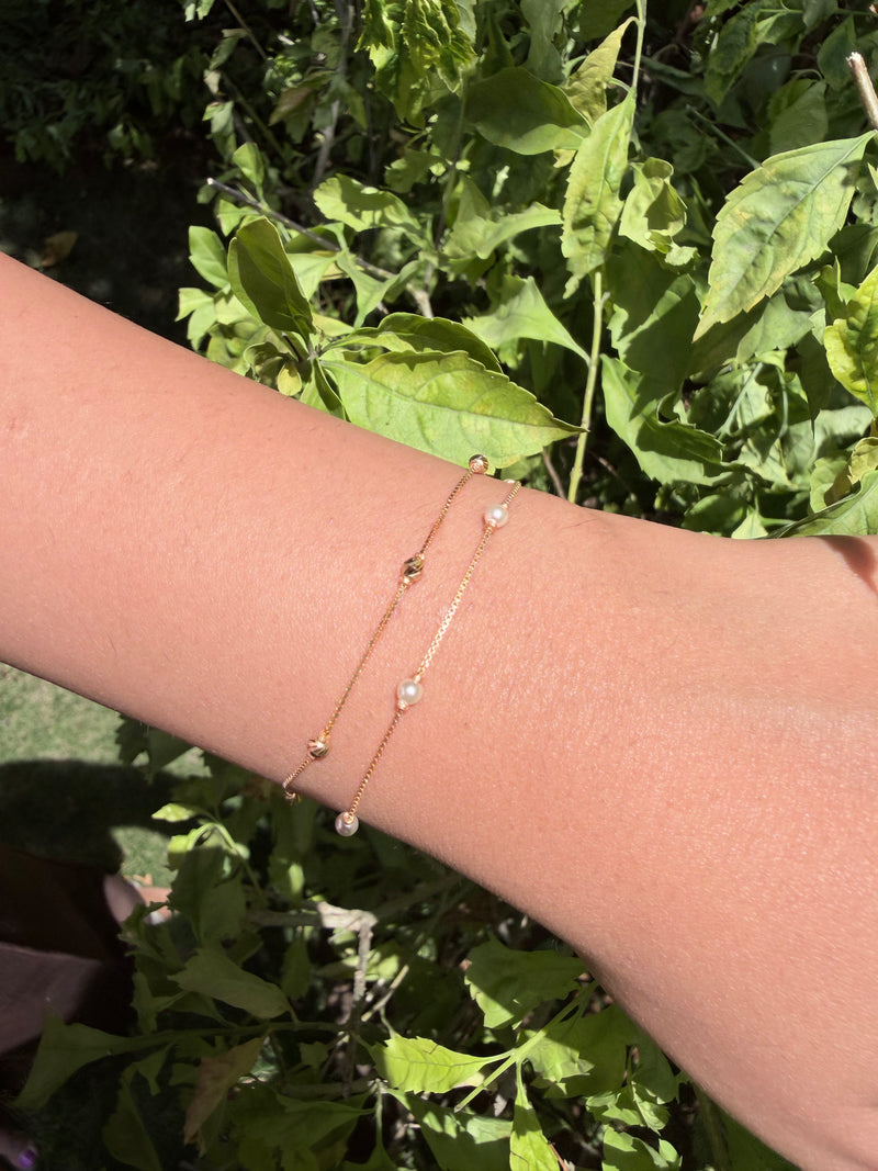 A close-up of a person's wrist wearing two delicate gold chain bracelets with small clear beads, set against a background of green outdoor foliage.