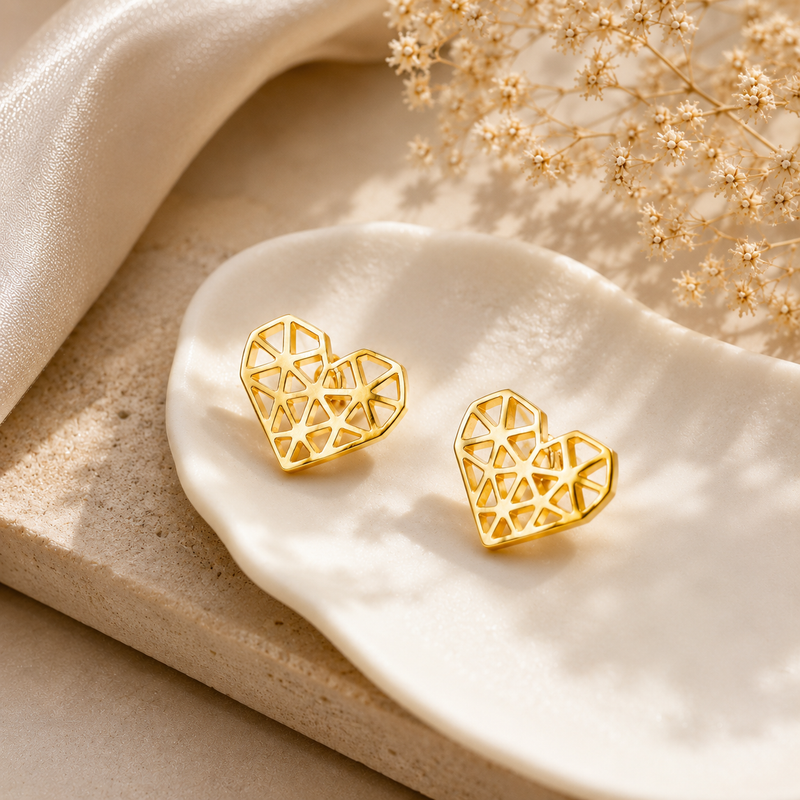 A pair of gold geometric heart stud earrings displayed on a white ceramic dish with neutral beige props and dried flowers.