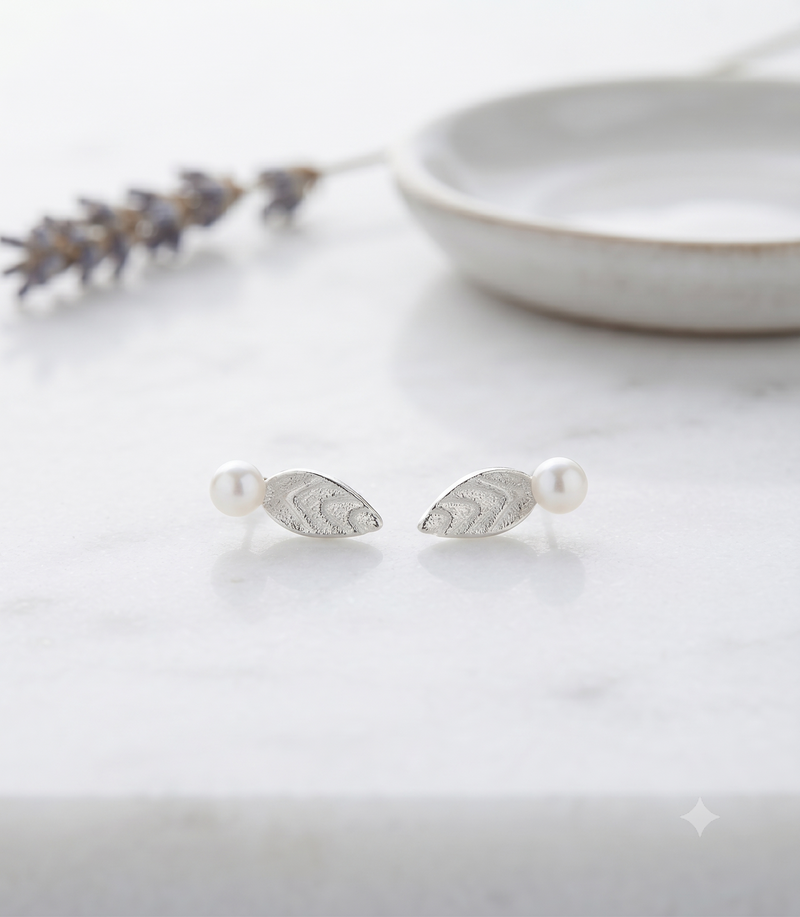A pair of silver leaf-shaped stud earrings with white pearl accents, displayed on a light marble surface with a blurred ceramic dish and lavender sprig in the background.