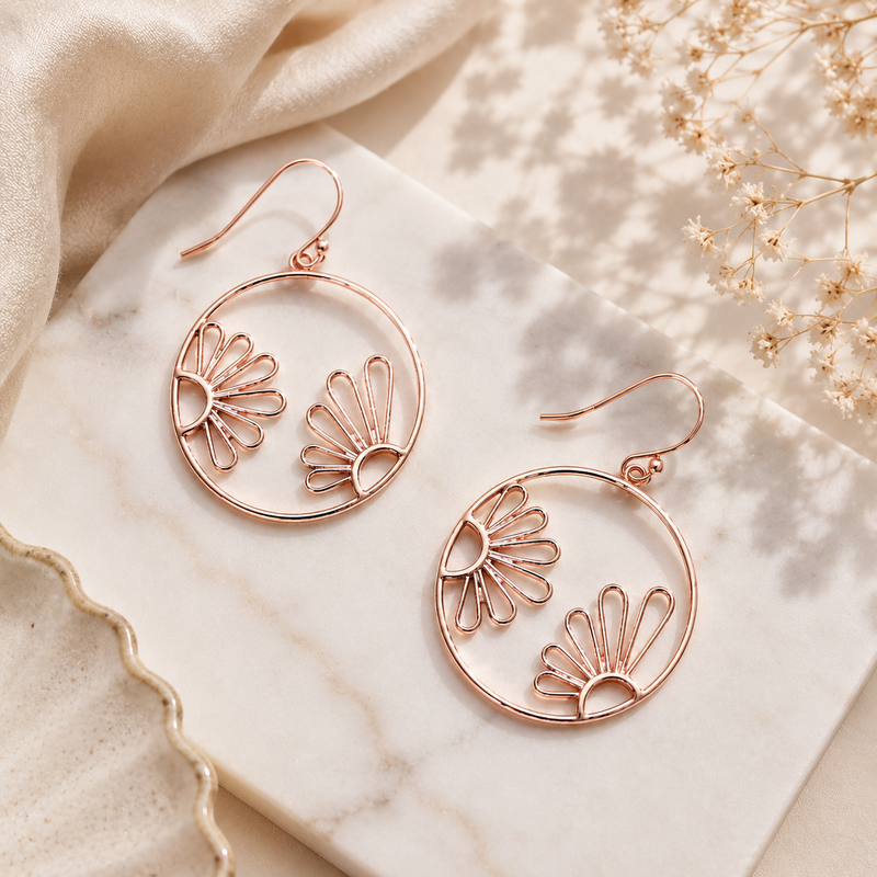 A pair of rose gold hoop drop earrings with a cutout floral design, displayed on a white marble tray with beige fabric and dried flowers in the background.
