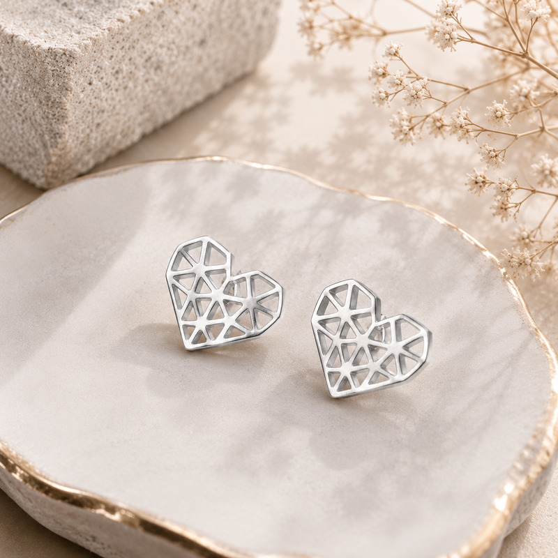 A pair of silver geometric heart stud earrings displayed on a white ceramic dish with gold trim, styled with a textured stone and dried baby's breath flowers.