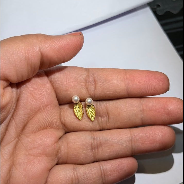 A pair of gold leaf stud earrings with small white pearl-like studs, shown resting on an open hand.