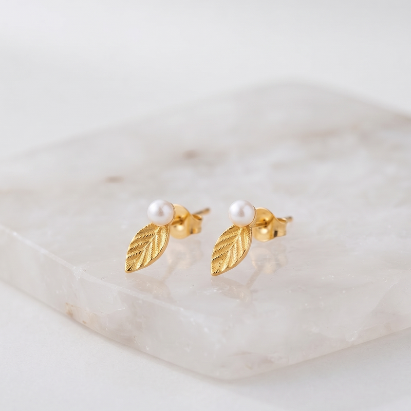 A pair of gold leaf stud earrings with a small white pearl at the center, displayed on a light marble block against a soft white background.