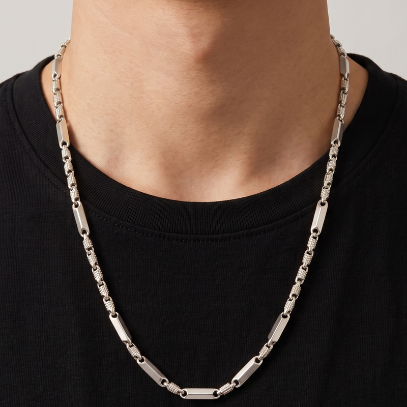 A close-up of a man's neck and chest wearing a silver-toned metal link chain necklace over a black t-shirt, against a light gray background.
