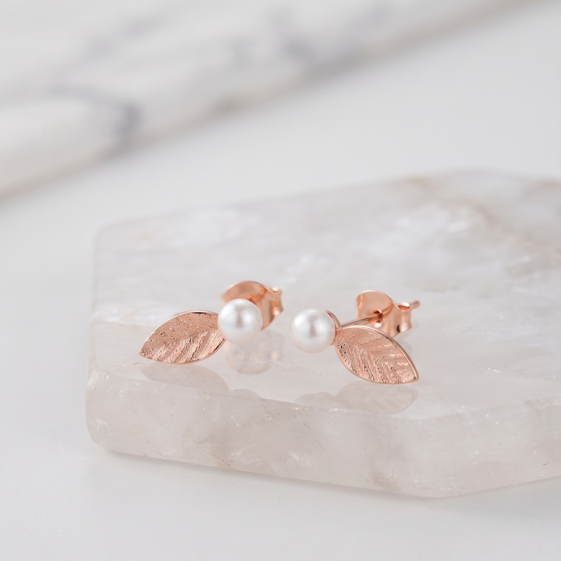 A pair of rose gold leaf stud earrings with white pearl centers, displayed on a polished white crystal block.