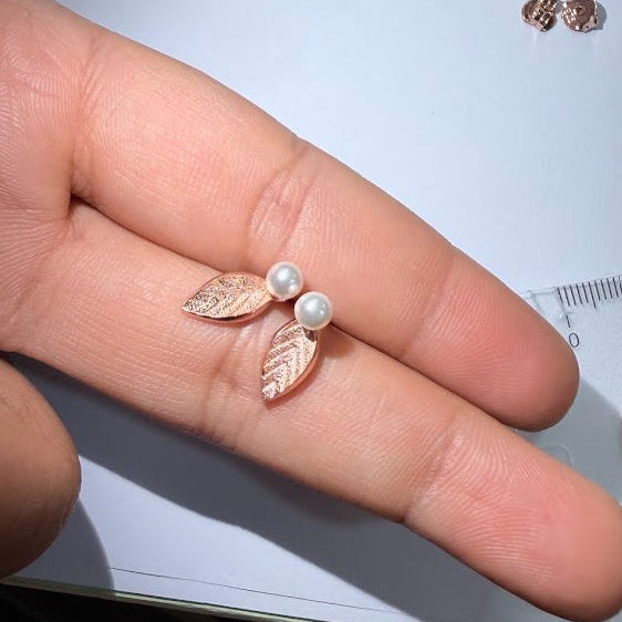 A pair of rose gold leaf stud earrings with white faux pearls, shown resting on a person's fingers for scale against a white background.