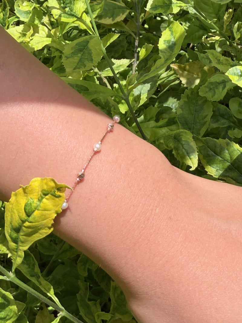 A delicate beaded chain bracelet with small white pearls and a tiny metallic bead, shown worn on a person's wrist against a background of green garden foliage.