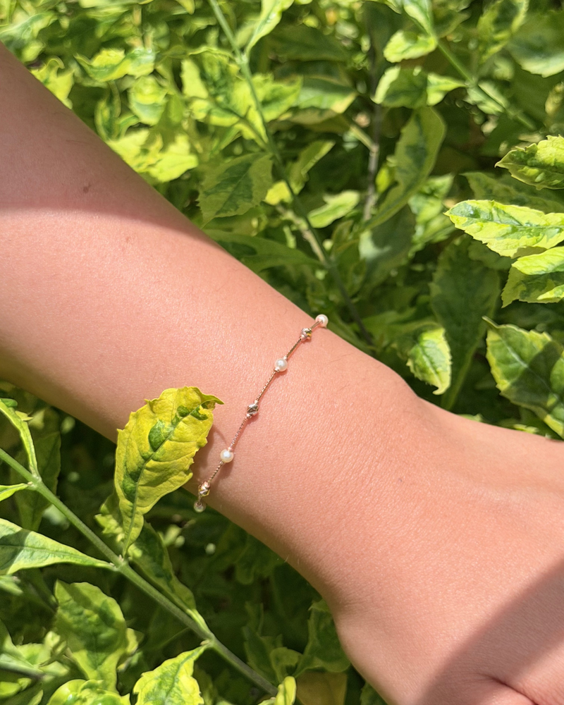 A delicate rose gold chain bracelet with small white pearl-like beads worn on a person's wrist, set against a background of green leaves.