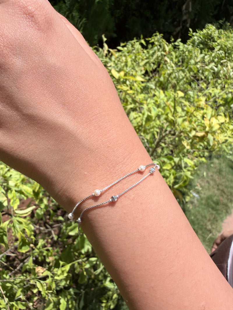 A close-up of a person's wrist wearing a delicate double-strand silver chain bracelet with small white beads and tiny star charms, set against a blurred green garden background.