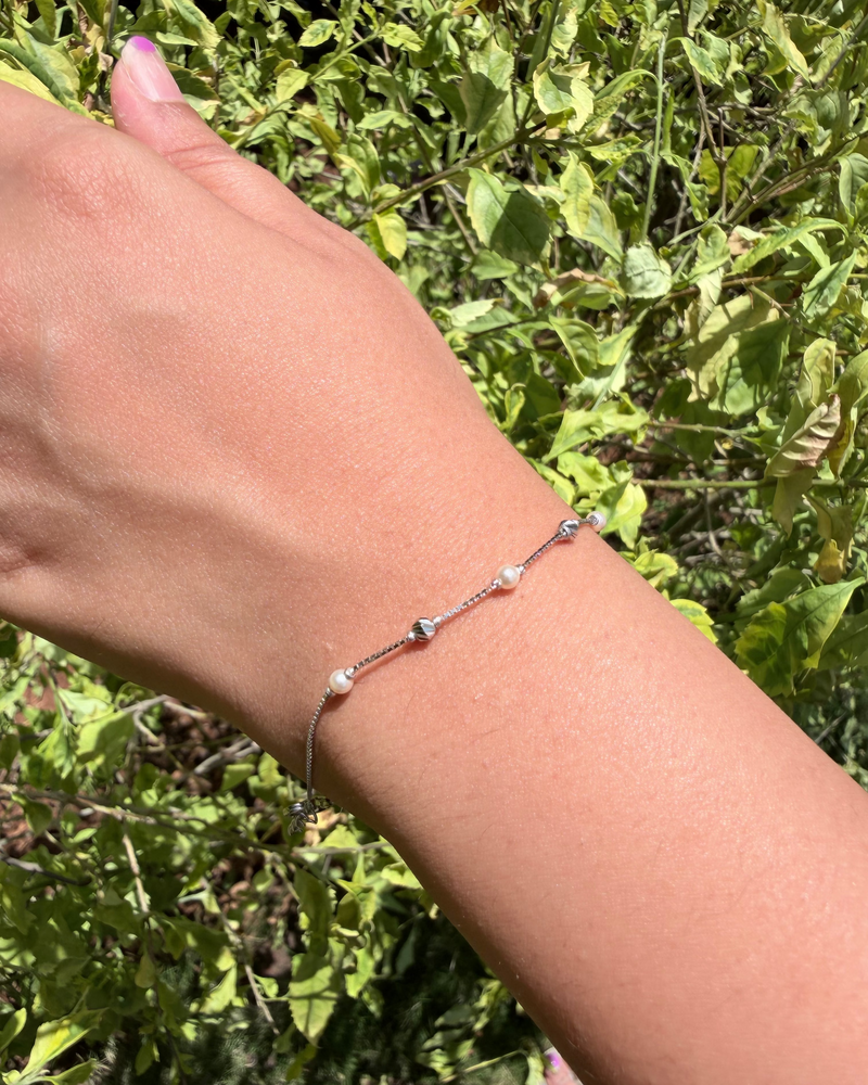 A delicate silver chain bracelet with small white pearl-like beads and a tiny silver heart charm, shown worn on a person's wrist against a background of green outdoor foliage.