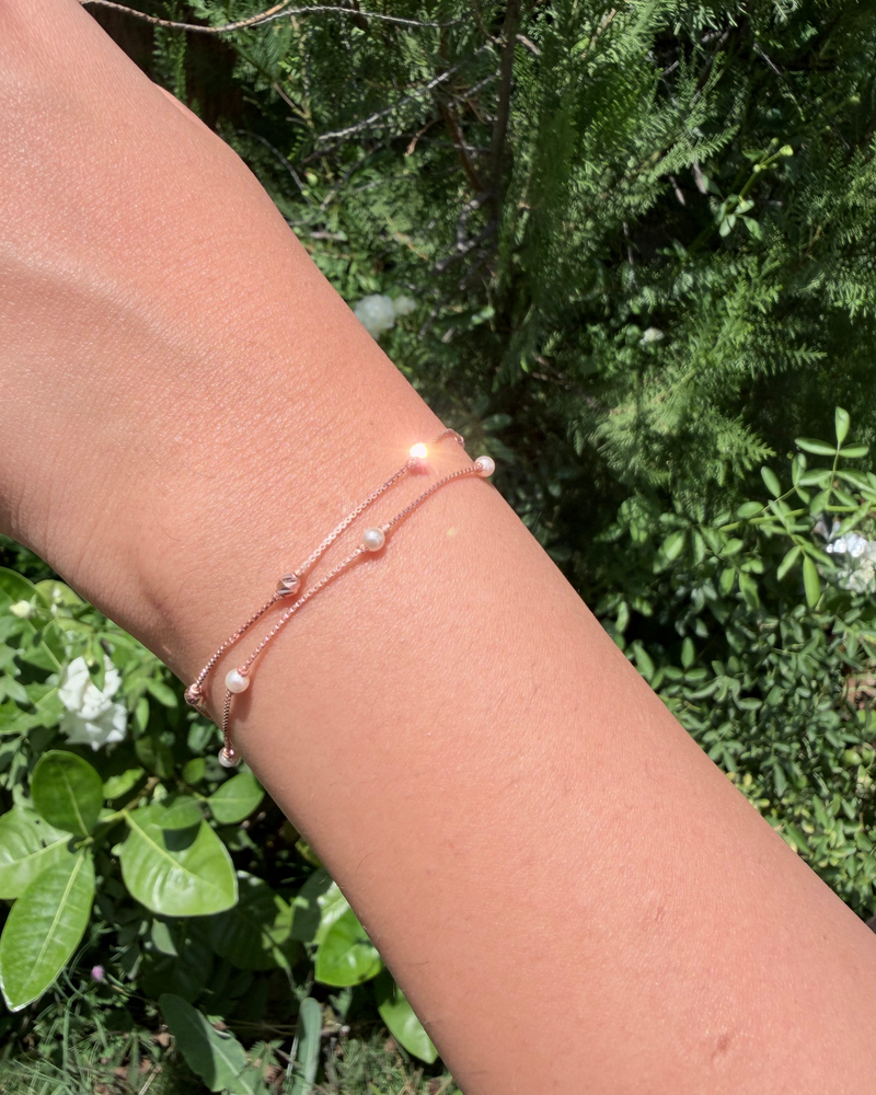 A close-up of a person's wrist wearing two delicate rose gold chain bracelets with small white pearl-like beads, set against a background of green garden foliage.