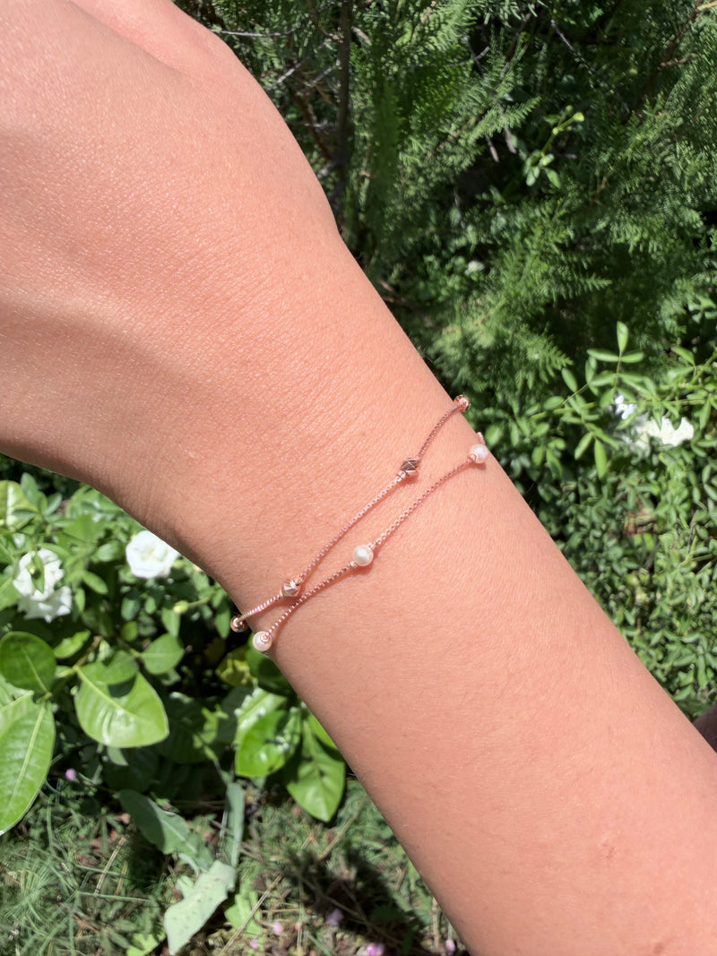 A close-up of a person's wrist wearing two delicate rose gold bracelets with small white pearl-like beads and tiny clear stones, set against a background of green garden foliage.