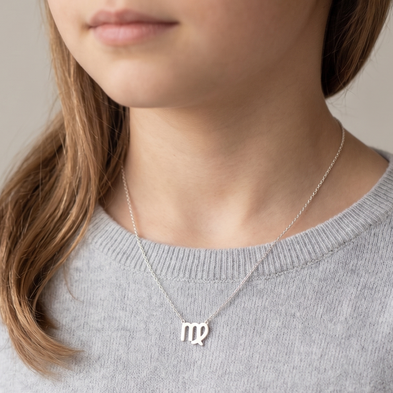 A close-up of a young girl wearing a delicate silver necklace with a 'mp' initial pendant, styled with a light gray knit sweater against a soft beige background.